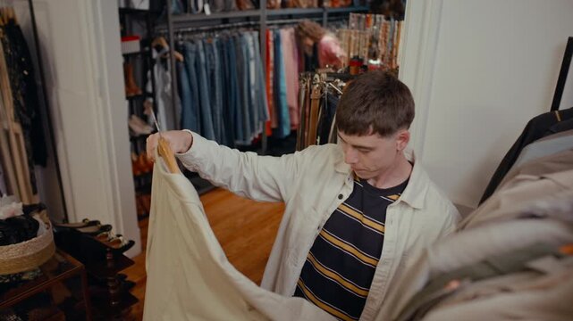 Young man examining beige jacket on hanger while browsing through clothing racks inside vintage thrift store