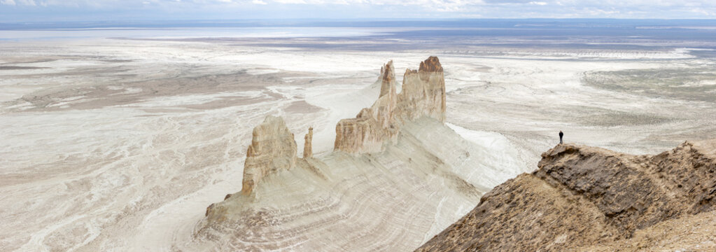 Aerial view of the dramatic Bozjyra rock formations rising from the vast desert landscape with a person standing on a cliff edge in Aktau, Mangystau Region, Kazakhstan.