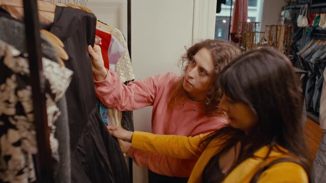Two young women smiling and discussing stylish outfits while browsing rack of secondhand clothes together in vintage thrift shop