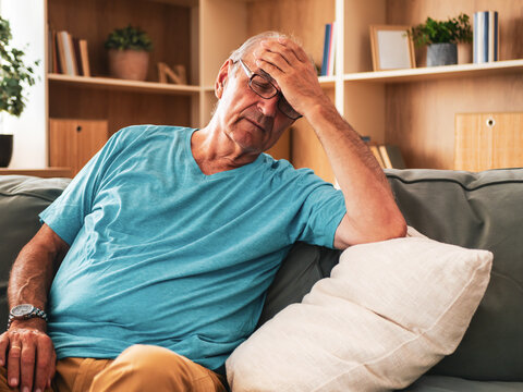 Elderly man sitting on sofa at home, suffering from headache, anxiety and stress, feeling tired and worried about his health.