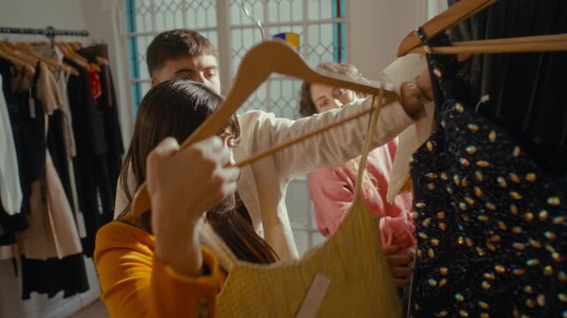 Young woman taking hanger with dress off rack, examining it and discussing with friends while choosing outfits together in cozy vintage thrift shop
