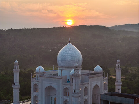 Aerial view of Budi Guna Mosque featuring a prominent white dome and minarets illuminated by the warm light of a sunset over the surrounding hills in Citeureup, West Java, Indonesia.