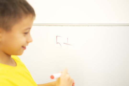 Close-up of a school age boy holding a red marker beside a handwritten number five and plus sign on a whiteboard during an individual educational activity.