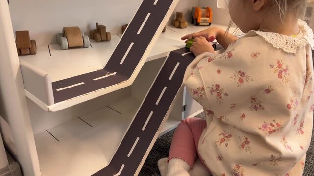A girl sends miniature cars down ramps on a wooden toy parking garage, imaginative play, childhood learning, motor skills.