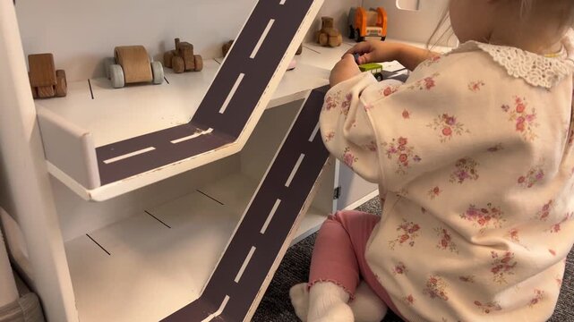 A girl moves small cars across the upper level of a wooden toy garage, creative play, childhood exploration, motor skills.