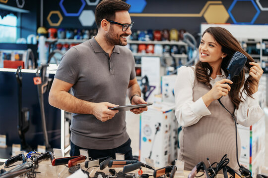 Retail store worker explaining product features on a digital tablet to an expectant mother testing a hair styling brush at an electronics market counter.