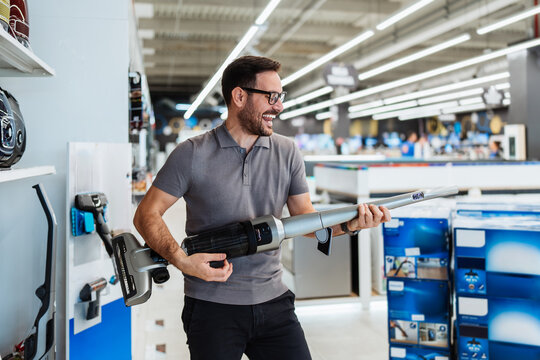 Happy male customer testing a modern home appliance device while browsing through a large retail shopping center aisles filled with household electronics.
