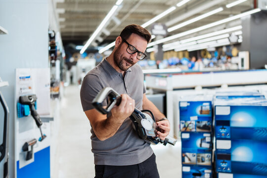 Happy male customer testing a modern home appliance device while browsing through a large retail shopping center aisles filled with household electronics.