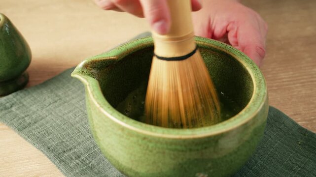 Whisking green matcha tea with traditional bamboo whisk chasen in ceramic bowl chawan to create foam. Preparing authentic Japanese matcha tea, healthy wellness beverage and detox drink.