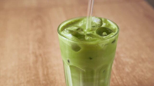 Stirring cold iced matcha green tea latte with glass straw in tall glass filled with ice cubes on wooden table. Iced matcha latte, healthy detox superfood beverage for daily summer wellness routine.