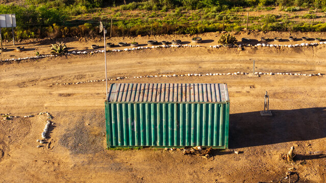 Aerial view of a green shipping container situated in a dry, dirt-covered lot with scattered tires and sparse vegetation in Ladismith, Western Cape, South Africa.
