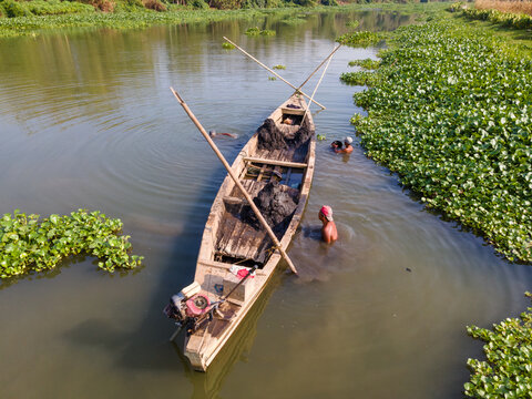 Aerial view of a wooden boat carrying sand on the Kalirajut river, with men working in the water surrounded by dense green water hyacinth in Patikraja, Central Java, Indonesia.