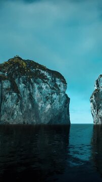 Two large rocky cliffs stand tall by a calm body of water. The scene captures a quiet moment with clear blue skies above. The sunlight reflects off the water, creating a natural view.