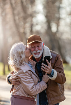 Happy senior woman kissing her smiling partner on the cheek while he takes a smartphone selfie in a sunny park, capturing a romantic moment of love and affection