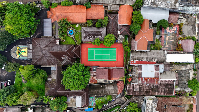 Aerial view of a tennis court surrounded by buildings with terracotta roofs, lush green trees, and a small circular fountain in Sukabumi, West Java, Indonesia.
