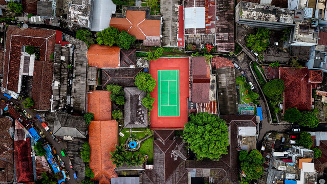 Aerial view of a bright green tennis court surrounded by dense urban rooftops and lush trees in Sukabumi, West Java, Indonesia.