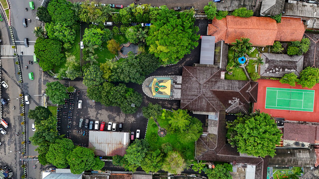 Aerial view of a building complex featuring a tennis court, lush green trees, a parking area with vehicles, and a distinctive roof structure in Sukabumi, West Java, Indonesia.