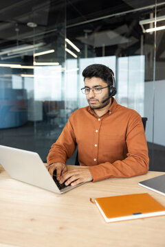 Young man wearing a headset and glasses providing remote customer support. Working on a laptop at a wooden desk in a modern office environment. Demonstrating professional assistance and communication