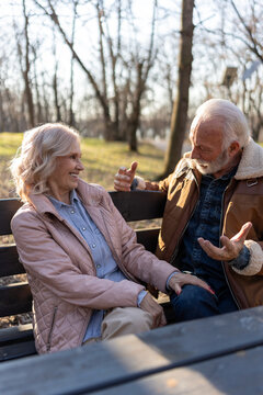 Senior couple sitting on a park bench, smiling and talking face to face while enjoying a relaxed conversation and quality time together on a sunny day