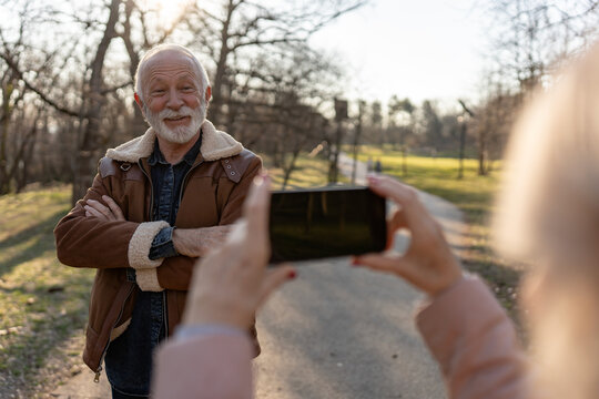 Happy senior man with white beard smiling and posing for a smartphone photo taken by a woman outdoors in a sunny park, enjoying a relaxed moment together