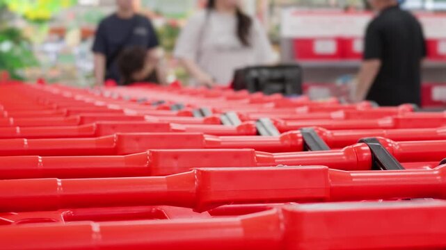 Close-up of a long row of nested red shopping carts with blurred figures of customers walking in the background inside a large modern supermarket or hypermarket. Retail trade and consumerism concept.