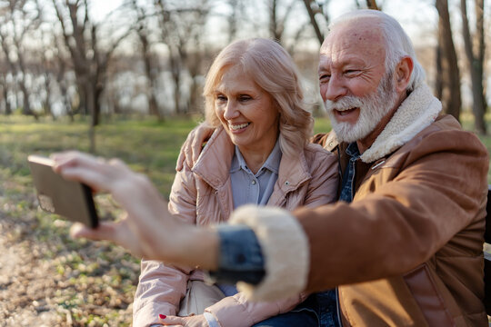 Happy senior couple sitting together on a park bench, laughing and taking a selfie with a smartphone, capturing joyful memories during a relaxing sunny day outdoors