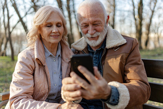 Happy senior couple using a smartphone together on a park bench, illustrating online banking, mobile financial services and digital independence during retirement