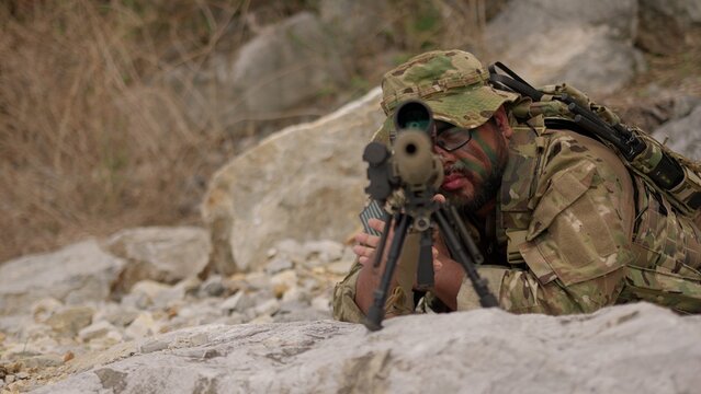 A special forces soldier in camouflage lies in a prone position, aiming his sniper rifle with a scope during a tactical surveillance mission in a rocky, arid terrain.
