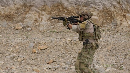 Fototapeta premium A special forces operator in full combat gear advances cautiously through a rocky, hostile environment while aiming his assault rifle on a tactical security patrol.