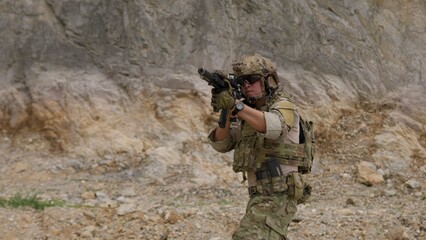 Fototapeta premium A special forces operator in full combat gear advances cautiously through a rocky, hostile environment while aiming his assault rifle on a tactical security patrol.