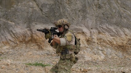 Fototapeta premium A special forces operator in full combat gear advances cautiously through a rocky, hostile environment while aiming his assault rifle on a tactical security patrol.