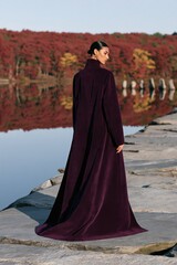 Fototapeta premium Woman wearing long purple velvet coat with flowing train walking on stone jetty over reflective lake with autumn trees for fashion editorial banner and poster