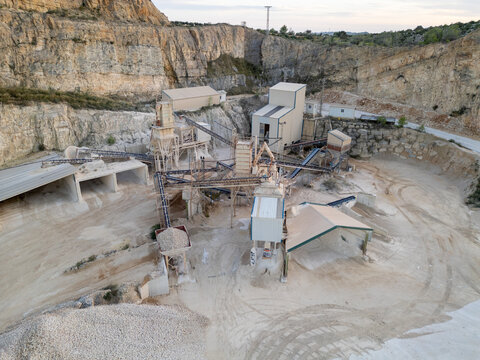 Aerial view of the stone quarry featuring industrial processing machinery, conveyor belts, and gravel piles set against a rocky cliff face in Teulada, Valencian Community, Spain.