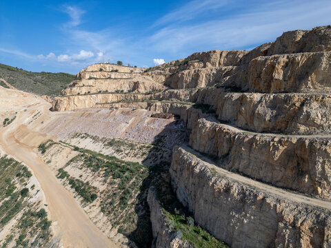 Aerial view of a large open-pit quarry with terraced rock faces, dirt access roads, and exposed stone layers under a clear blue sky in Teulada, Valencian Community, Spain.