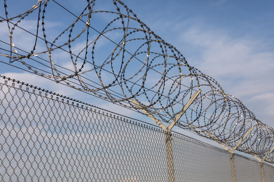 07 October 2026, Coiled razor wire sits atop a metal chain-link security fence against a blue sky.