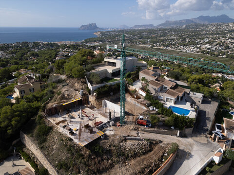 Aerial view of a construction site with a tall green crane, concrete foundations, and surrounding villas with swimming pools overlooking the coastline in Moraira, Valencian Community, Spain.