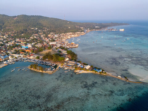 Aerial view of Karimun Jawa island featuring the coastal village, a busy harbor with small boats, turquoise shallow waters, and lush green hills in Karimunjawa, Central Java, Indonesia.