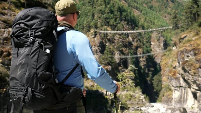 Trekker with large backpack admiring amazing himalayan landscape with long suspension bridge hillary spanning deep gorge on sunny day in everest region of nepal