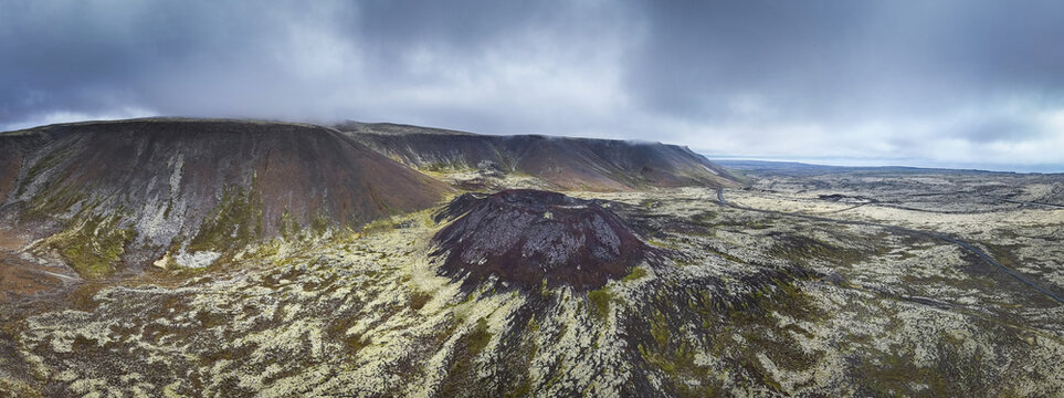 Aerial view of the volcanic Eldborg Crater surrounded by moss-covered lava fields and dramatic basaltic hills under a cloudy sky in Eldborg Crater, Borgarbyggd, Iceland.