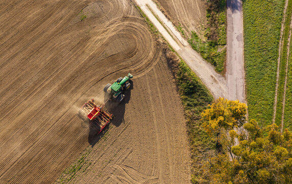 Aerial view of a green tractor pulling a red agricultural seeder across a large, freshly plowed brown field next to a dirt road and green vegetation in Pomeranian Voivodeship, Poland.