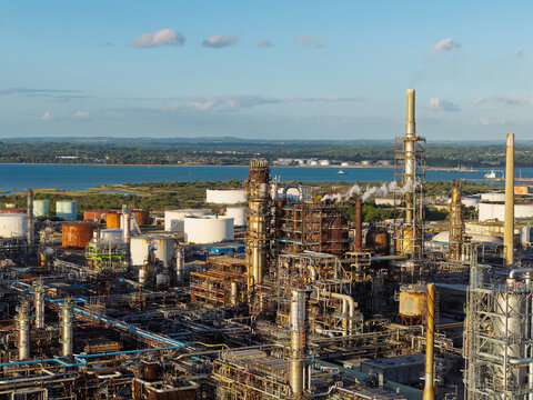 Aerial view of an oil refinery with industrial towers, storage tanks, and pipelines under a clear blue sky in the United Kingdom.