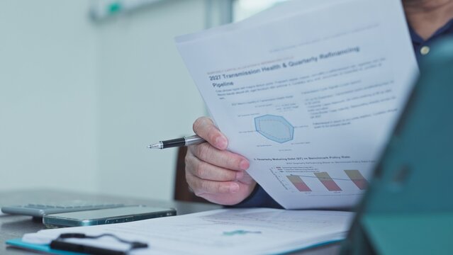 Businessman holding pen reviews printed financial report sheets showing debt refinancing data, investment charts, economic graphs seated beside tablet computer at workplace desk indoors.