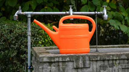bright orange plastic watering can resting on a concrete basin beneath outdoor water taps © Flowal93