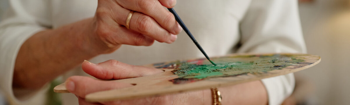 Close up of senior woman hands holding wooden palette and mixing paint with brush during art workshop. Visual for senior hobby clubs and creative therapy marketing
