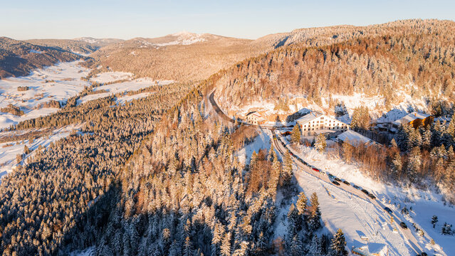 Aerial view of Col de la Faucille in winter, featuring a winding road through snow-covered pine forests and mountain slopes in Gex, Auvergne-Rhone-Alpes, France.