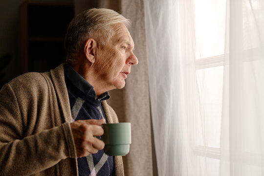Senior Caucasian man holding cup and looking through window thoughtfully. Image suitable for retirement lifestyle, elderly mental wellbeing campaigns, nursing home advertising