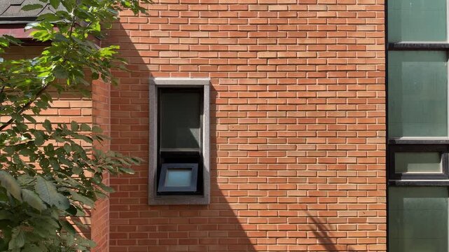Sunlit Red Brick Wall with a Small Open Window, Geometric Shadow Patterns and Green Leaves, Textured Architecture Facade in Natural Daylight