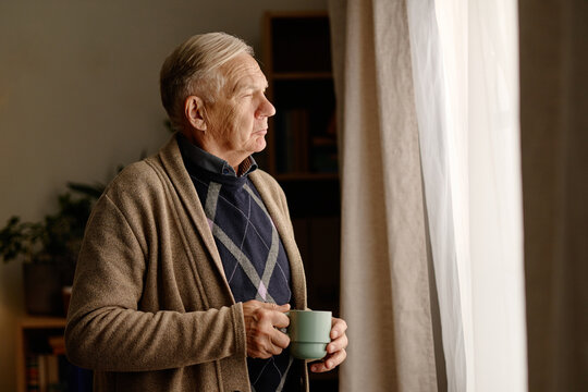 Senior Caucasian man standing near window holding cup, looking outside thoughtfully. Ideal for retirement living, senior mental health, healthcare campaigns