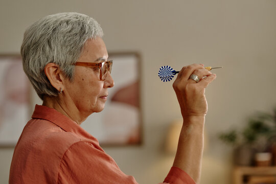 Senior Asian woman aiming dart during recreation in community club. Visual represents active aging and mental focus, ideal for senior lifestyle marketing and elderly wellness campaigns