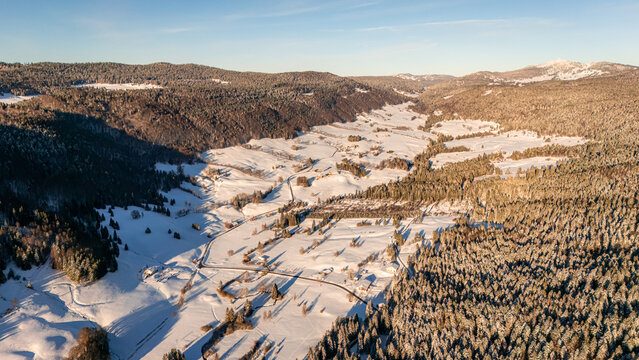 Aerial view of the French Jura in winter with a snow-covered valley, dense pine forests, and a winding road under golden hour light
